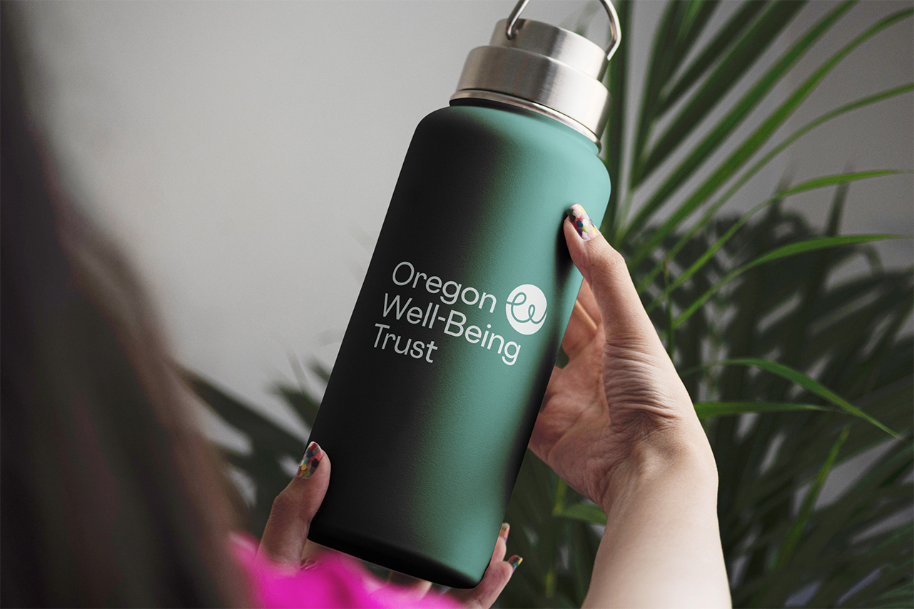 A close-up photograph of a hand holding a dark green water bottle with the Oregon Well-Being Trust's new logo printed on the front.