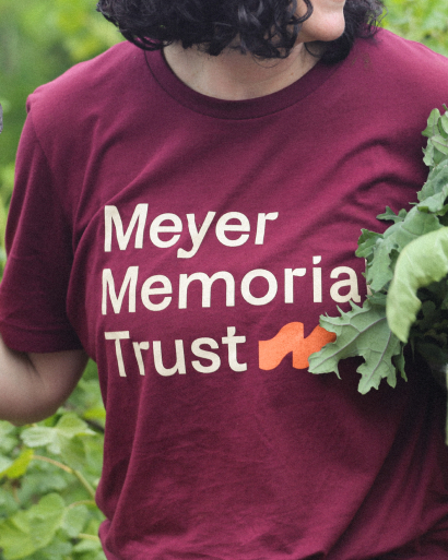 Person wearing a maroon Meyer Memorial Trust shirt holds fresh greens in a garden.