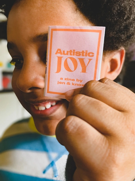 Smiling child holding up miniature zine cover that says Autistic Joy
