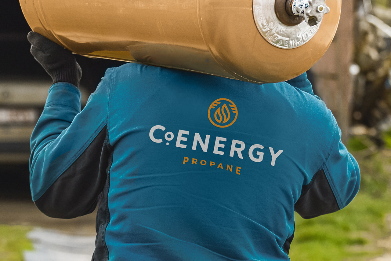 A photograph of a person carrying a propane tank, their back to the camera with the CoEnergy propane logo on the back of their jacket.
