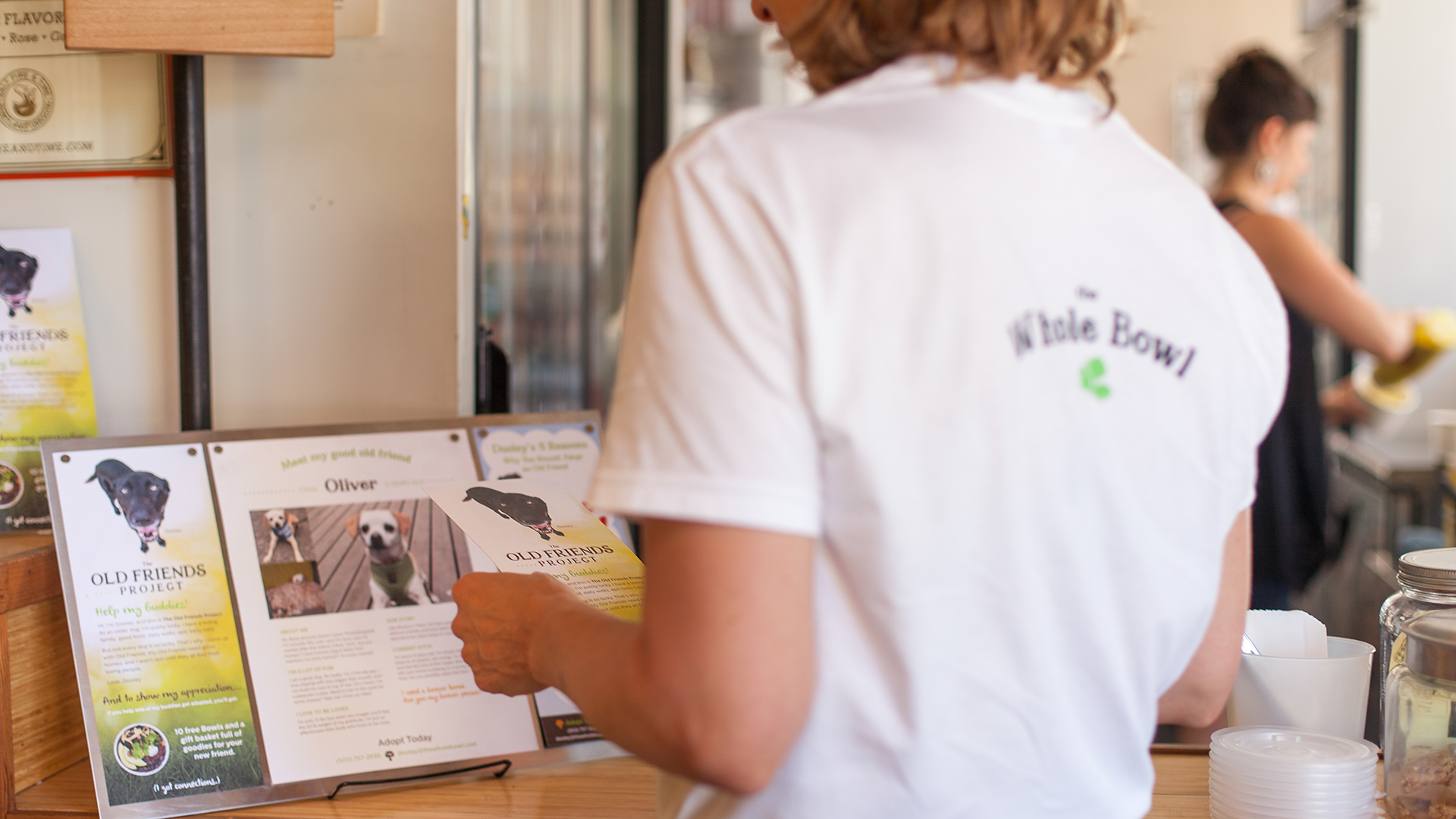 A photograph of a woman standing in a Whole Bowl restaurant wearing a white shirt with the Whole Bowl logo.